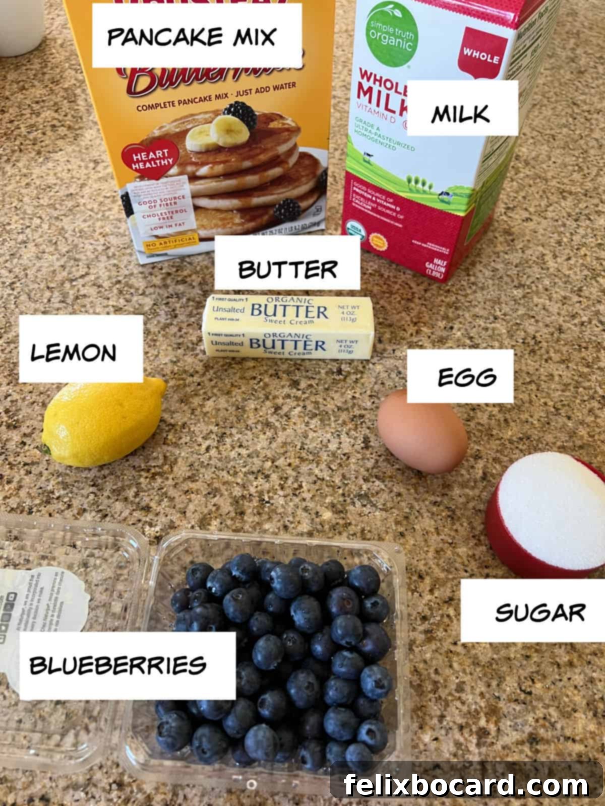 A beautiful flat lay of ingredients for pancake mix blueberry muffins, including pancake mix, milk, melted butter, fresh lemon, an egg, granulated sugar, and vibrant blueberries.