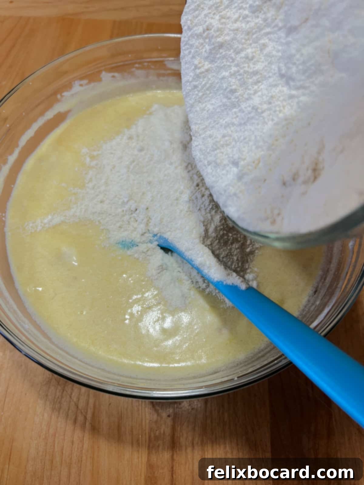 A top-down view of dry ingredients, including flour, baking powder, baking soda, and salt, being carefully poured into a large mixing bowl containing the wet banana and sour cream batter, captured at the crucial stage of combining the elements for muffin dough.