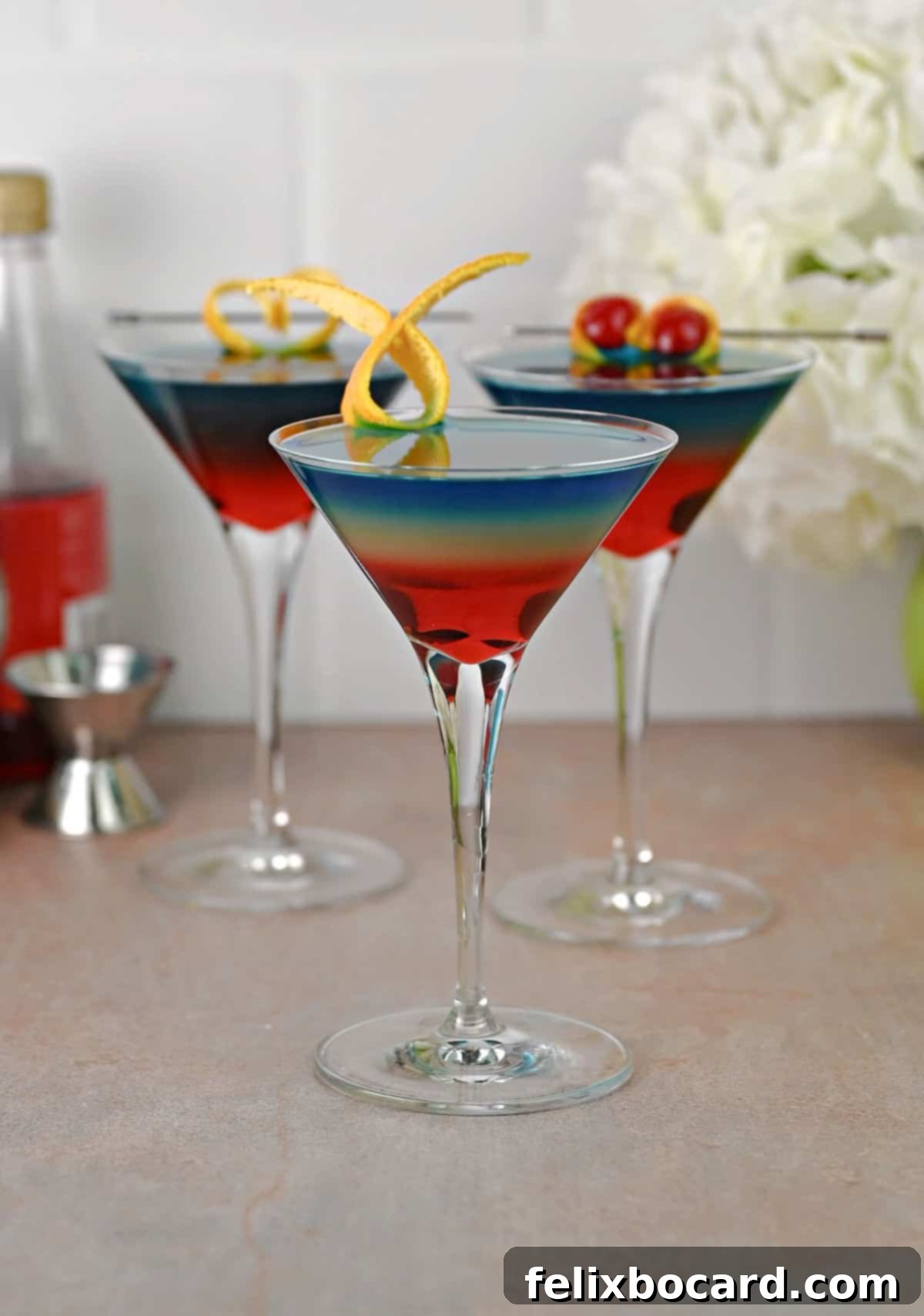 Three rainbow martini cocktails garnished with orange peels and cherries, showcasing distinct color layers.