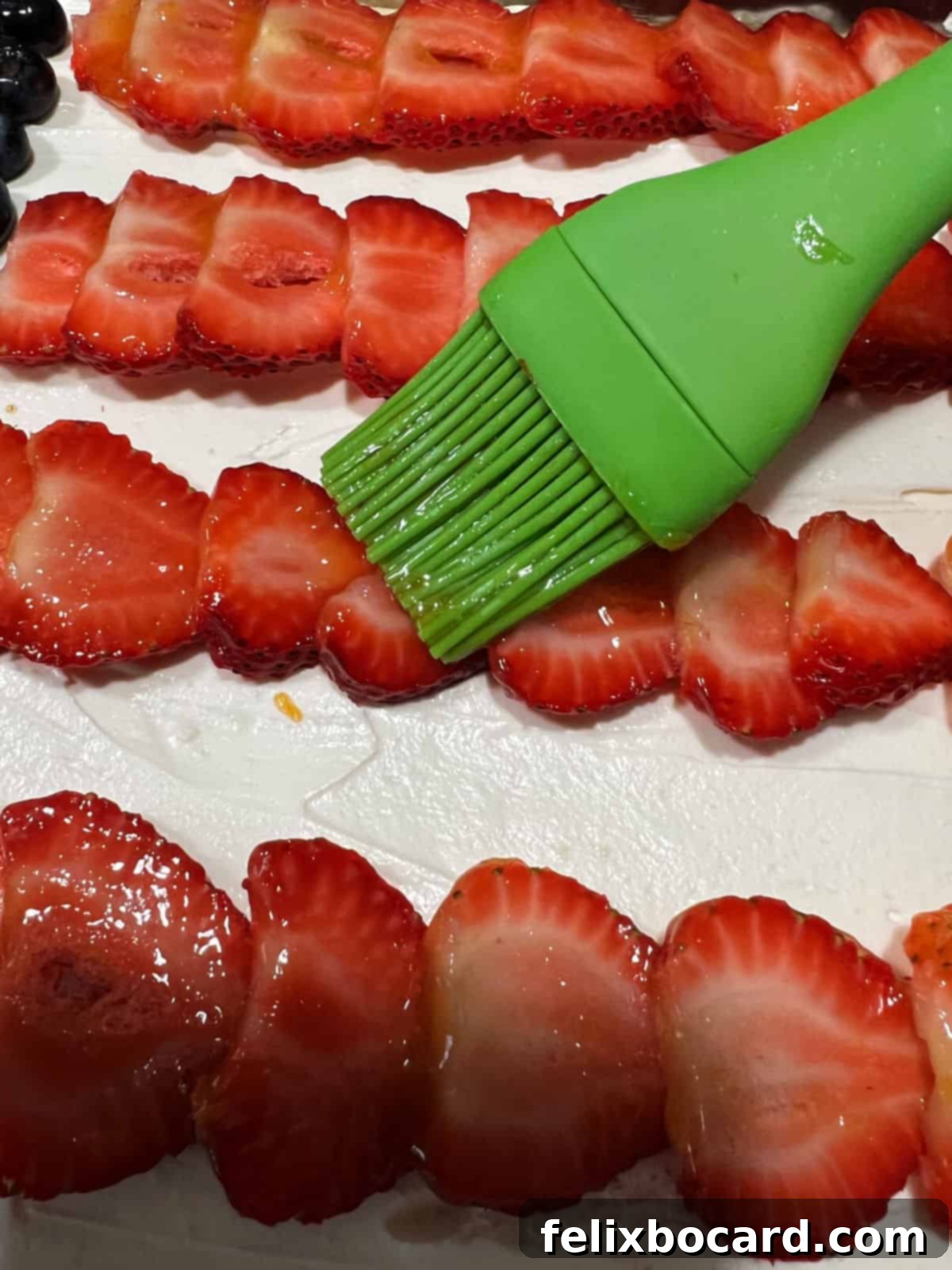 A pastry brush applying a light layer of apricot preserves to the fruit on an American Flag Fruit Pizza, adding a glossy finish.