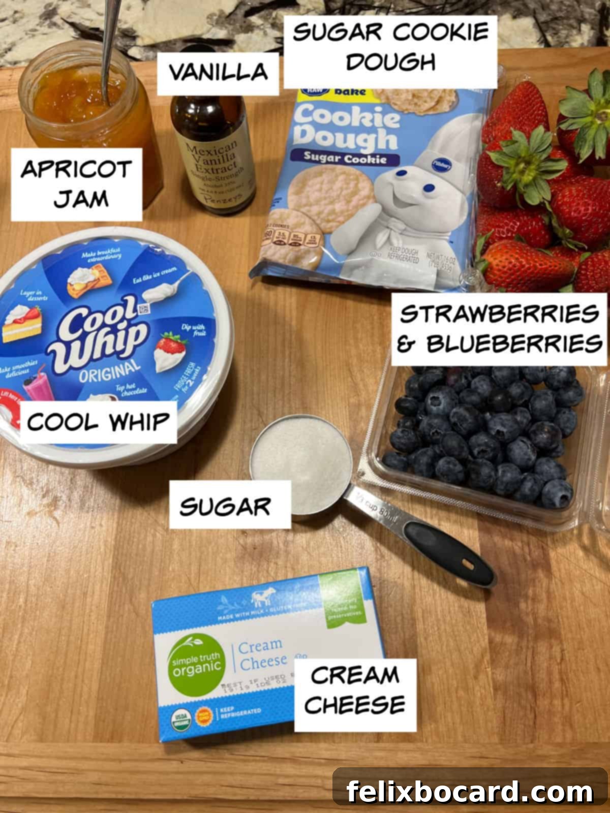 Ingredients laid out: cream cheese, sugar, Cool Whip, vanilla extract, a roll of sugar cookie dough, fresh strawberries and blueberries, and a jar of apricot jam.