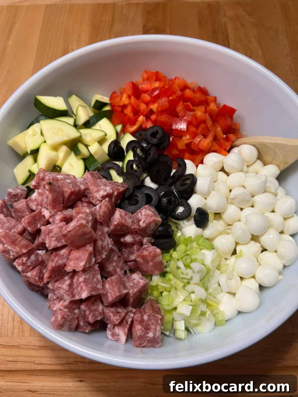 All ingredients added to bowl with pasta and dressing: salami, zucchini, bell pepper, olives, cheese, and green onions.
