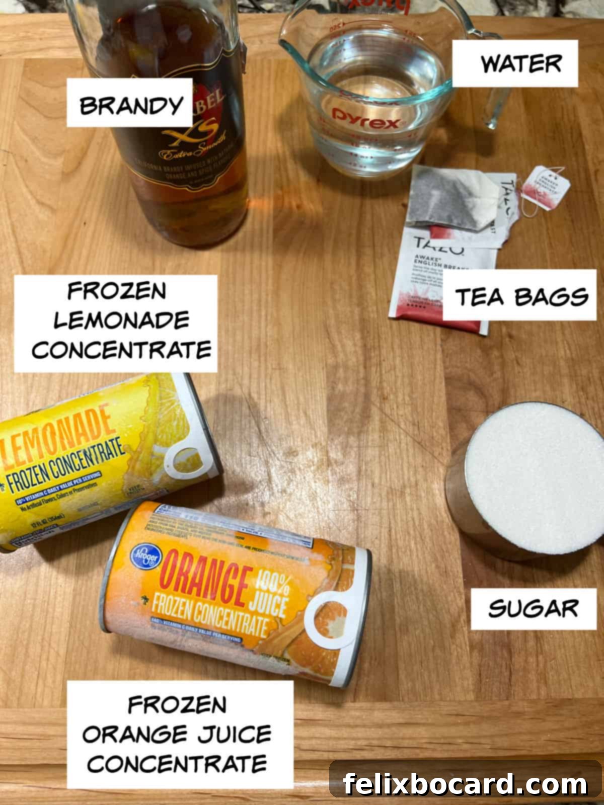 A flat lay photograph showcasing all the ingredients for Brandy Slush: a bottle of brandy, water in a glass, tea bags, granulated sugar, a can of orange juice concentrate, and a can of lemonade concentrate.