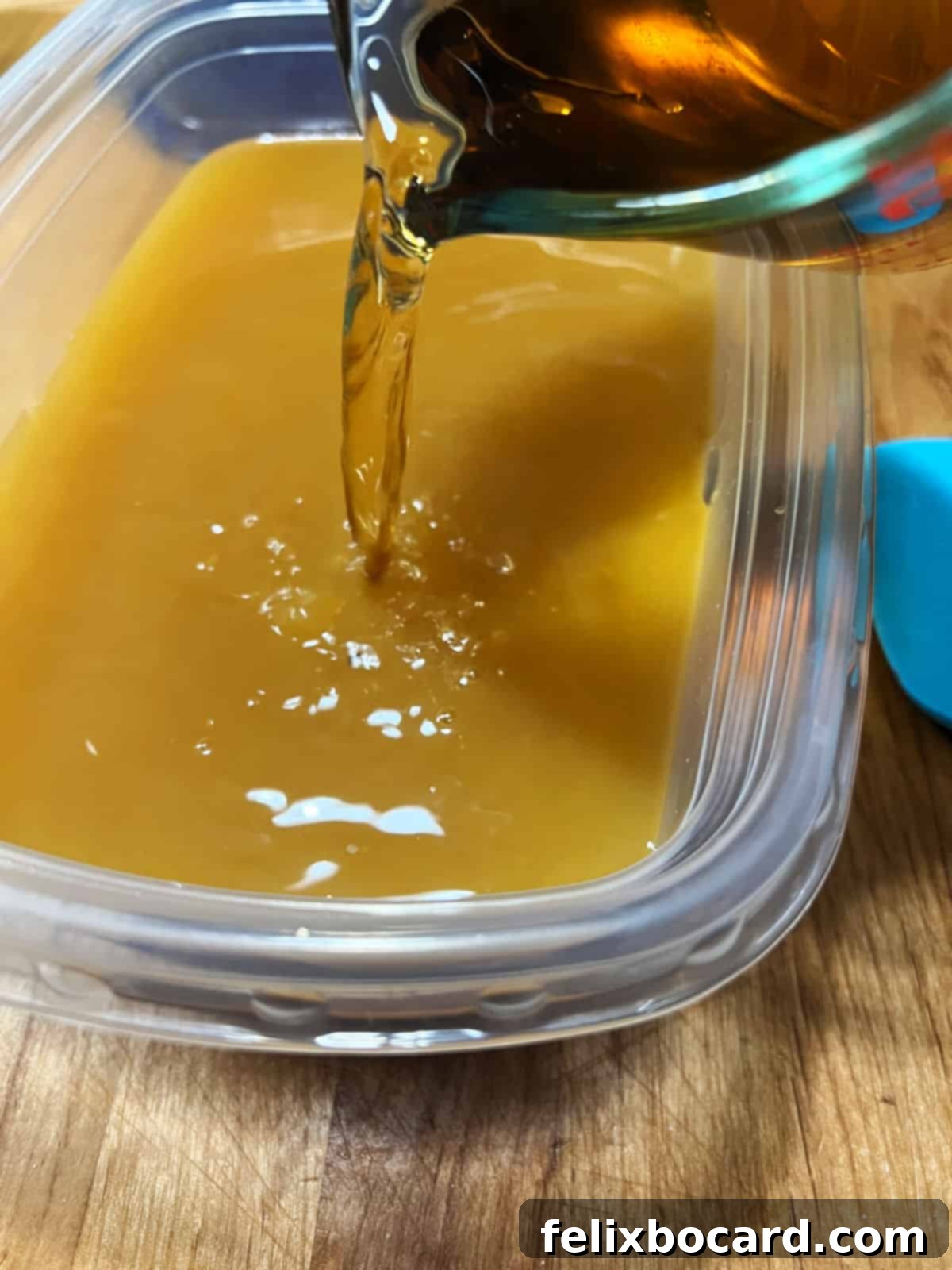 A hand pouring a stream of amber liquid, presumably brandy, from a bottle into a large clear bowl containing a mixture of other ingredients, illustrating the mixing process for Brandy Slush.
