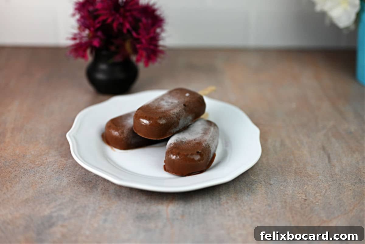 Three homemade fudgesicles thawing slightly on a rustic wooden plate, showcasing their creamy texture and rich chocolate color. Perfect for a summer treat.