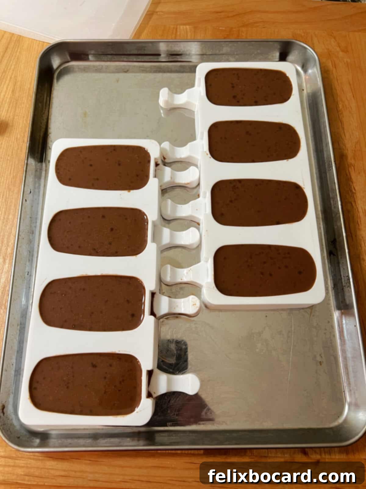 Several vibrant silicone popsicle molds are shown, each filled with the creamy chocolate fudgesicle mixture, ready for sticks.