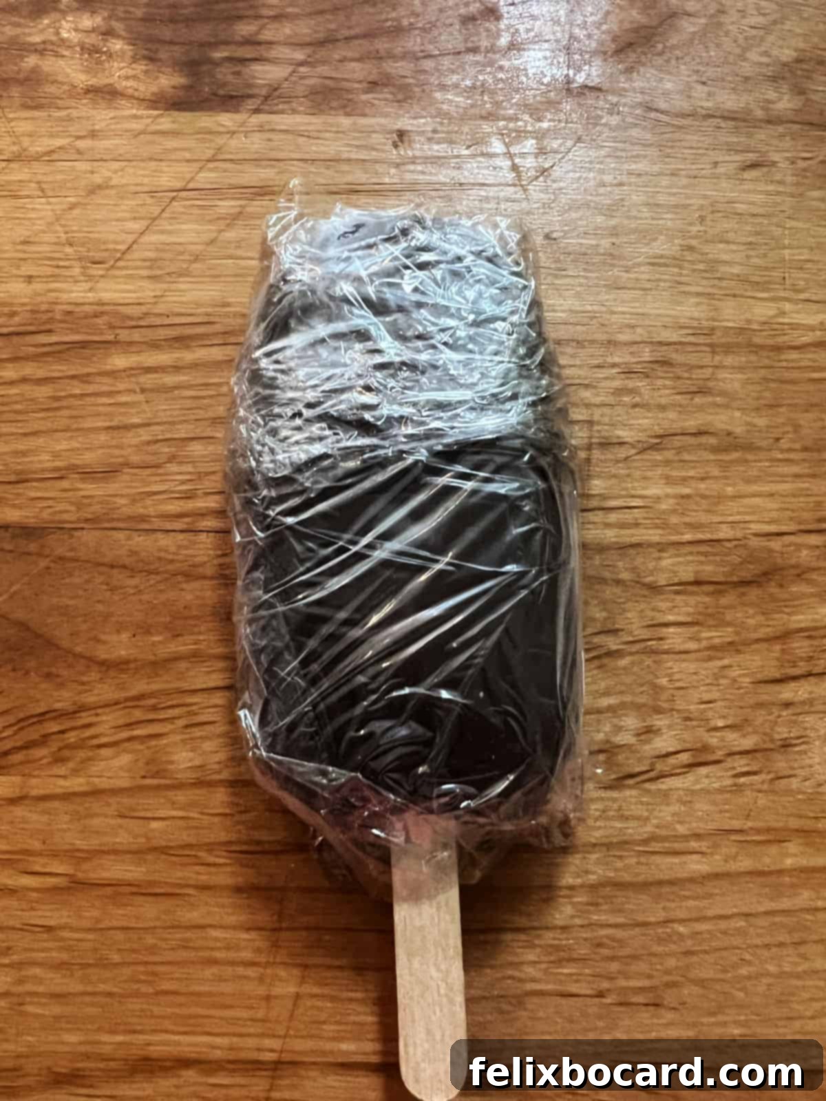 A homemade fudgesicle, perfectly removed from its mold and individually wrapped tightly in clear plastic wrap, ready for long-term freezer storage.
