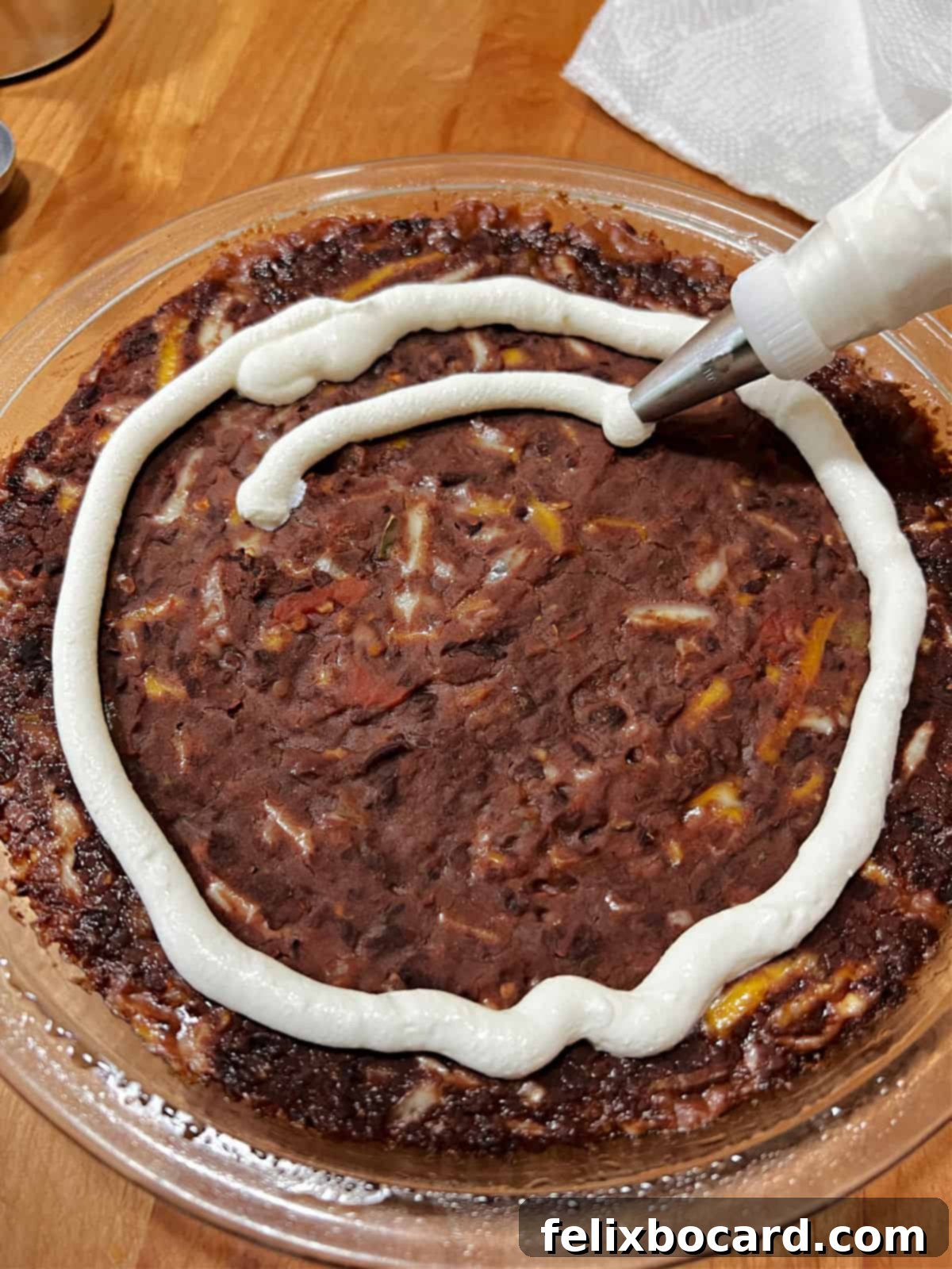 Using a pastry bag, concentric circles of sour cream are meticulously drawn on the hot surface of the black bean dip, forming the base of a spider web.