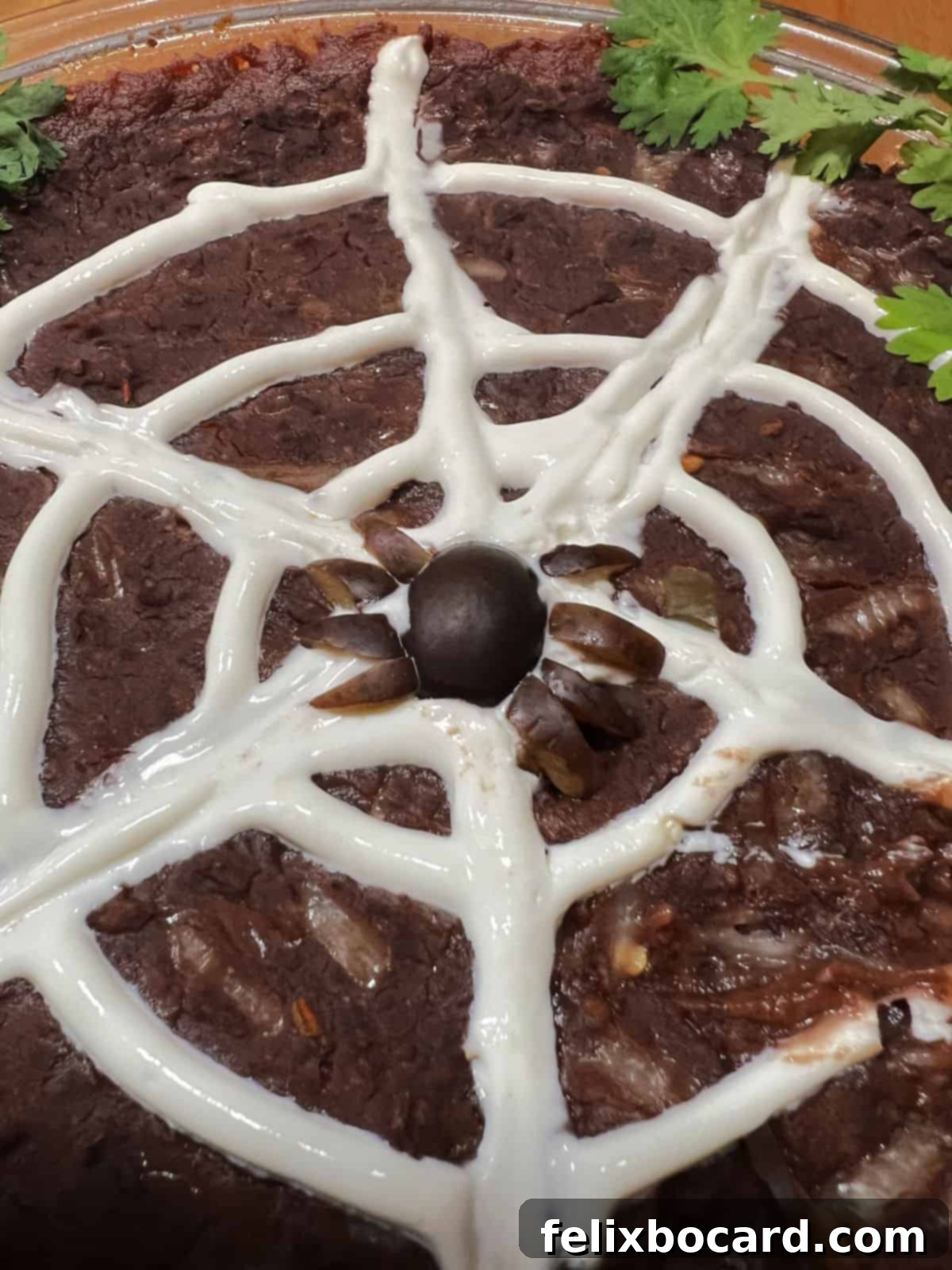An edible black olive spider dramatically perched on the sour cream web of the black bean dip, ready to be served at a Halloween gathering.