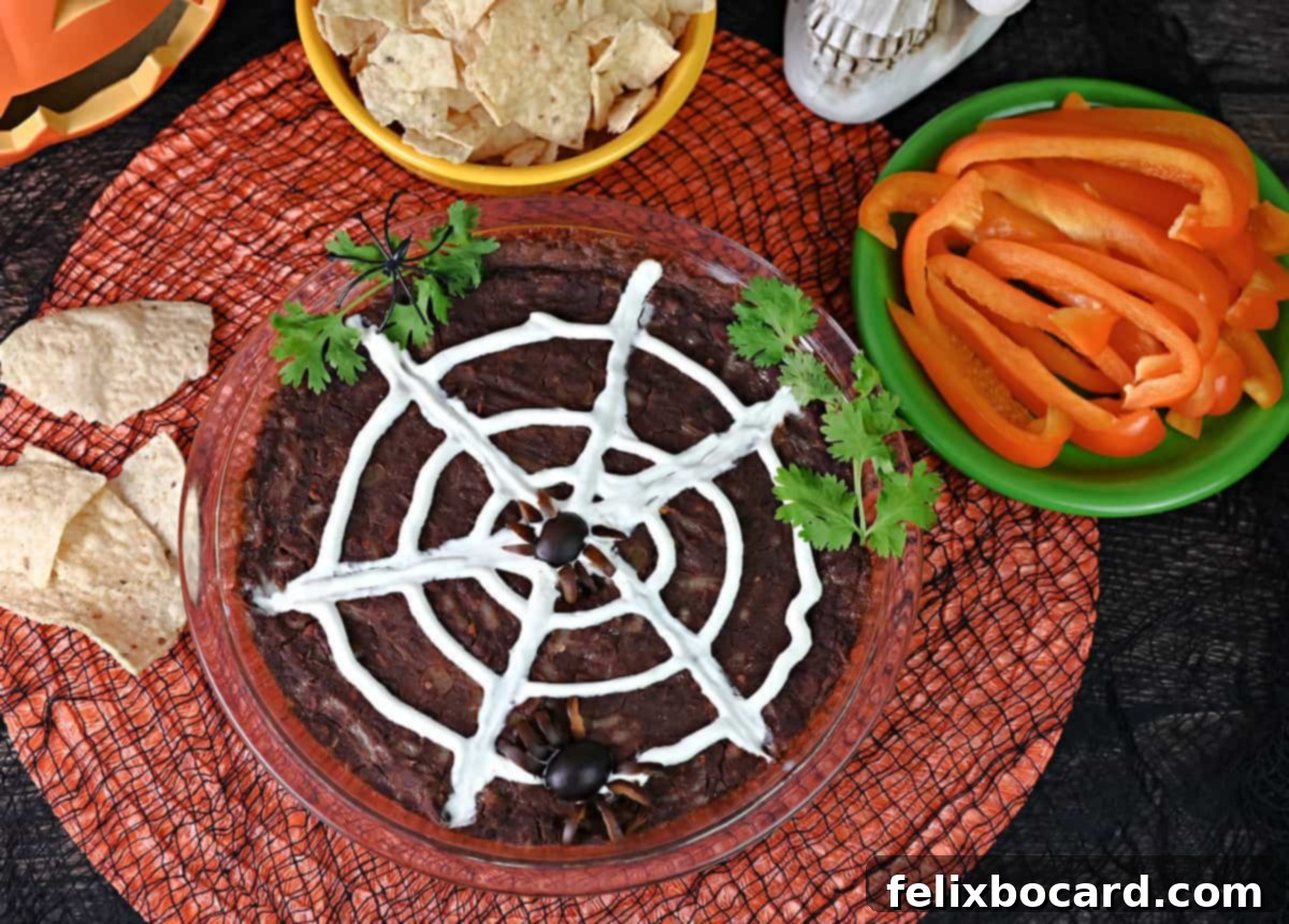The finished Spider Web Black Bean Dip, artfully decorated with sour cream and an olive spider, surrounded by a vibrant assortment of tortilla chips, bell pepper slices, and festive Halloween decor, ready for a party.