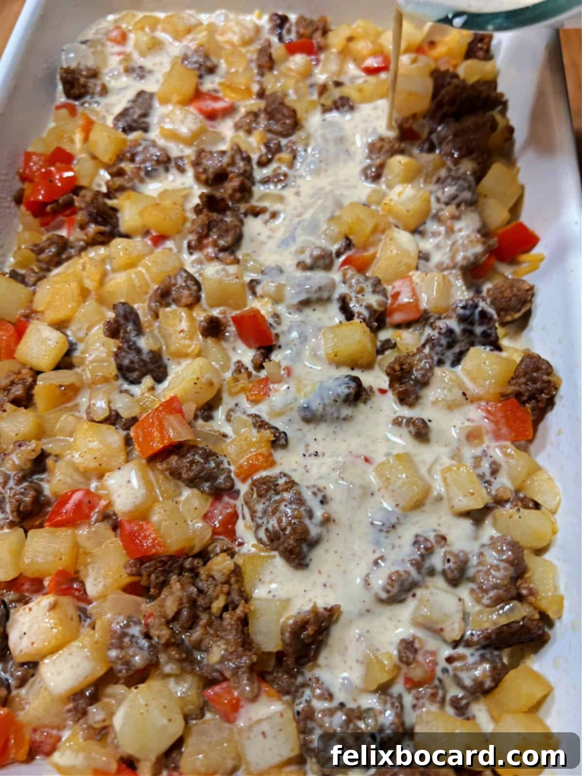 Hands pouring the whisked egg and cream mixture evenly over the first layer of chorizo, hash browns, and cheese in a baking dish.