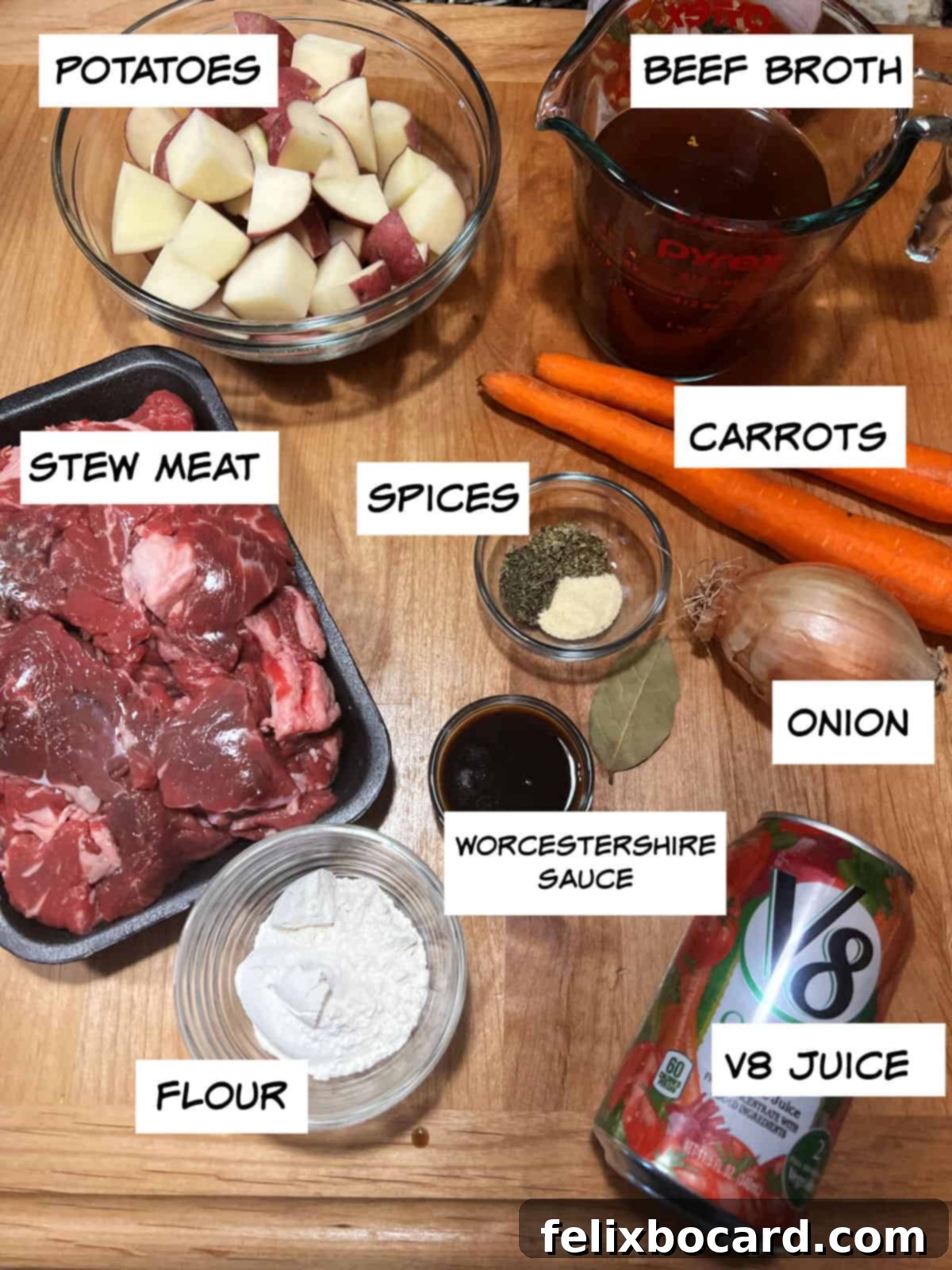 A vibrant collection of beef stew ingredients laid out on a countertop, including fresh potatoes, beef broth, crisp carrots, a yellow onion, raw stew meat, a bottle of V8 juice, all-purpose flour, Worcestershire sauce, and a selection of dried spices.