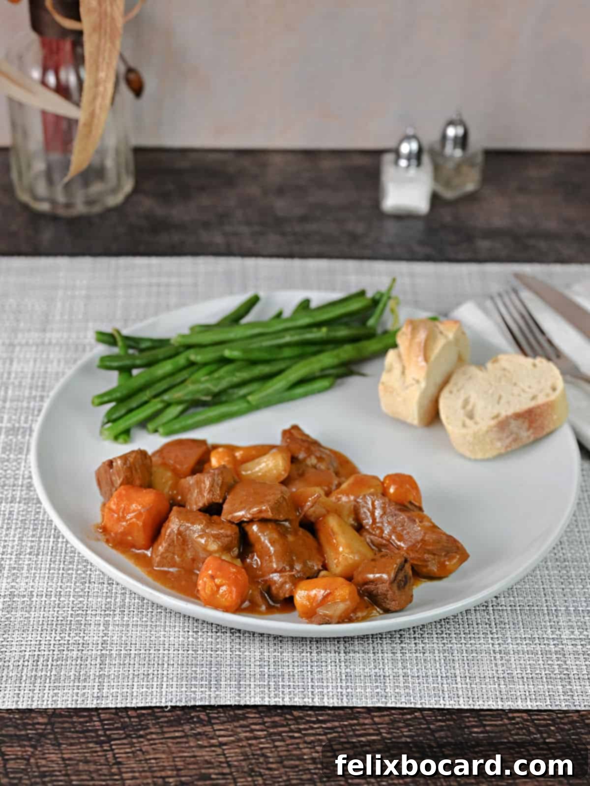 A generously plated portion of beef stew, artfully arranged with fresh green beans and a slice of rustic bread, all presented on a dark wooden surface.