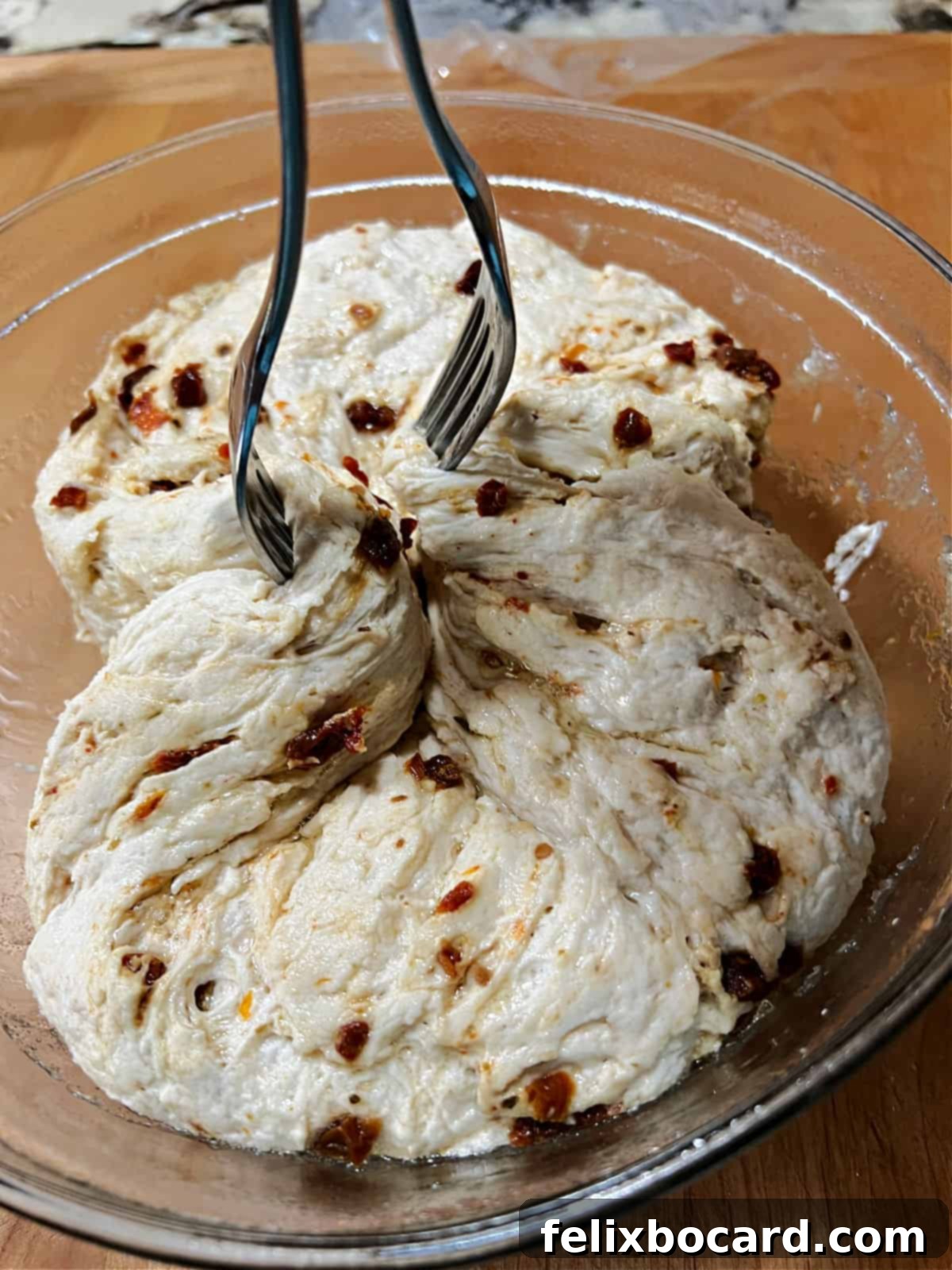 Simple Sun-Dried Tomato & Rosemary Focaccia 7 A baker using two forks to gently reshape and lift the focaccia dough in its bowl, preparing it for transfer.