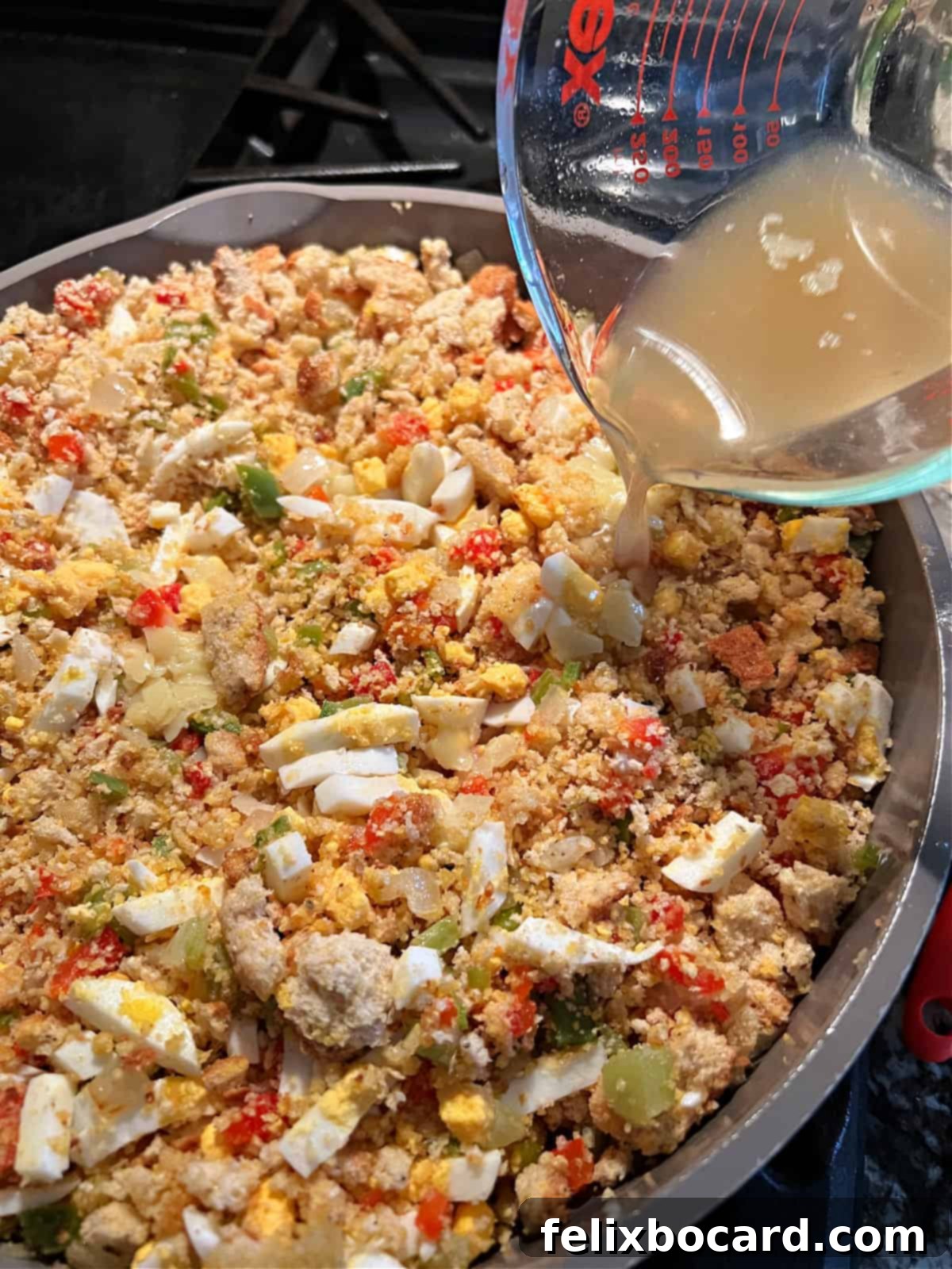 A large skillet containing the cornbread dressing mixture, with a stream of chicken broth being poured over it, ready for mixing.