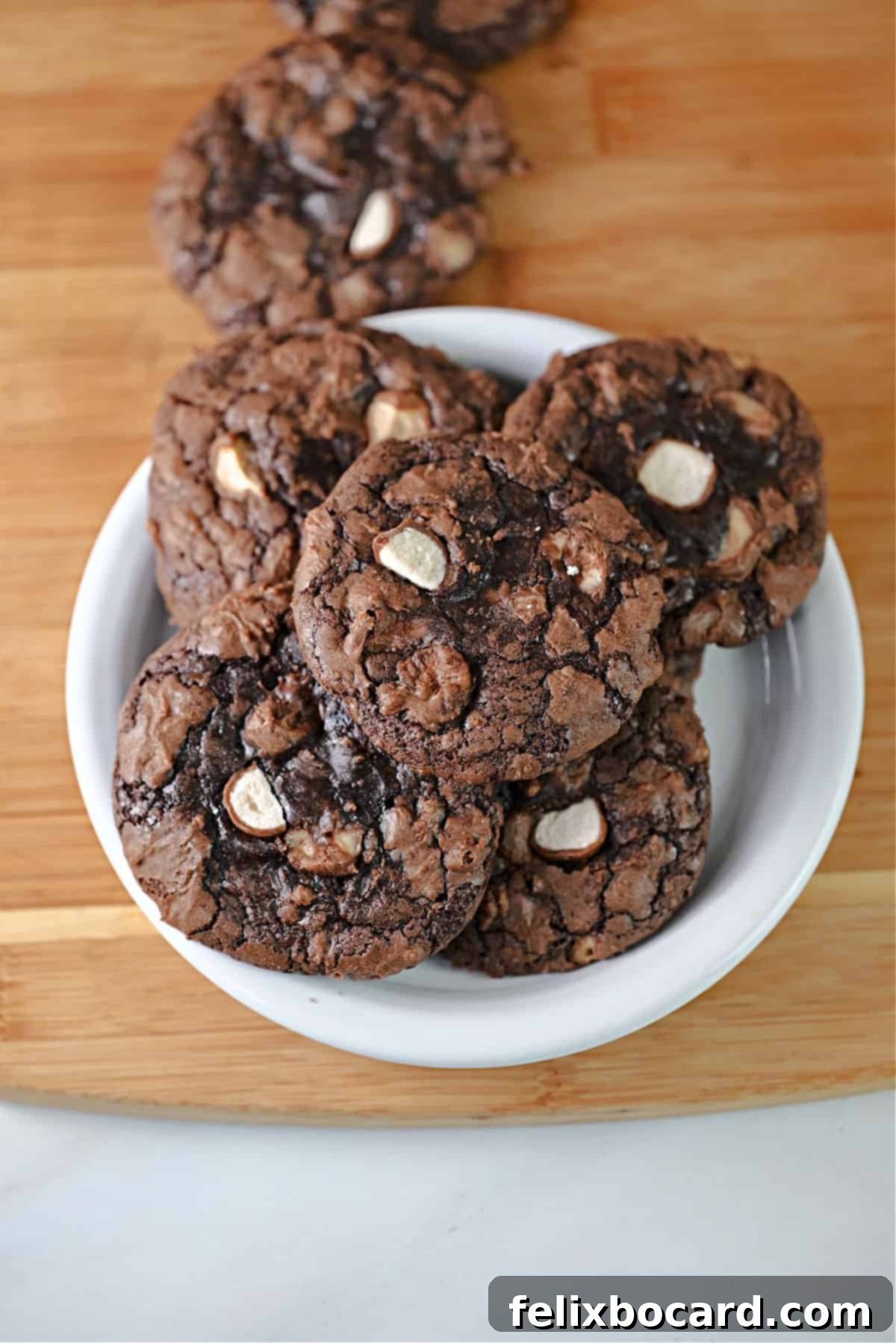 A small plate of chocolate malted milk balls cookies sitting on a cutting board with more cookies nearby.