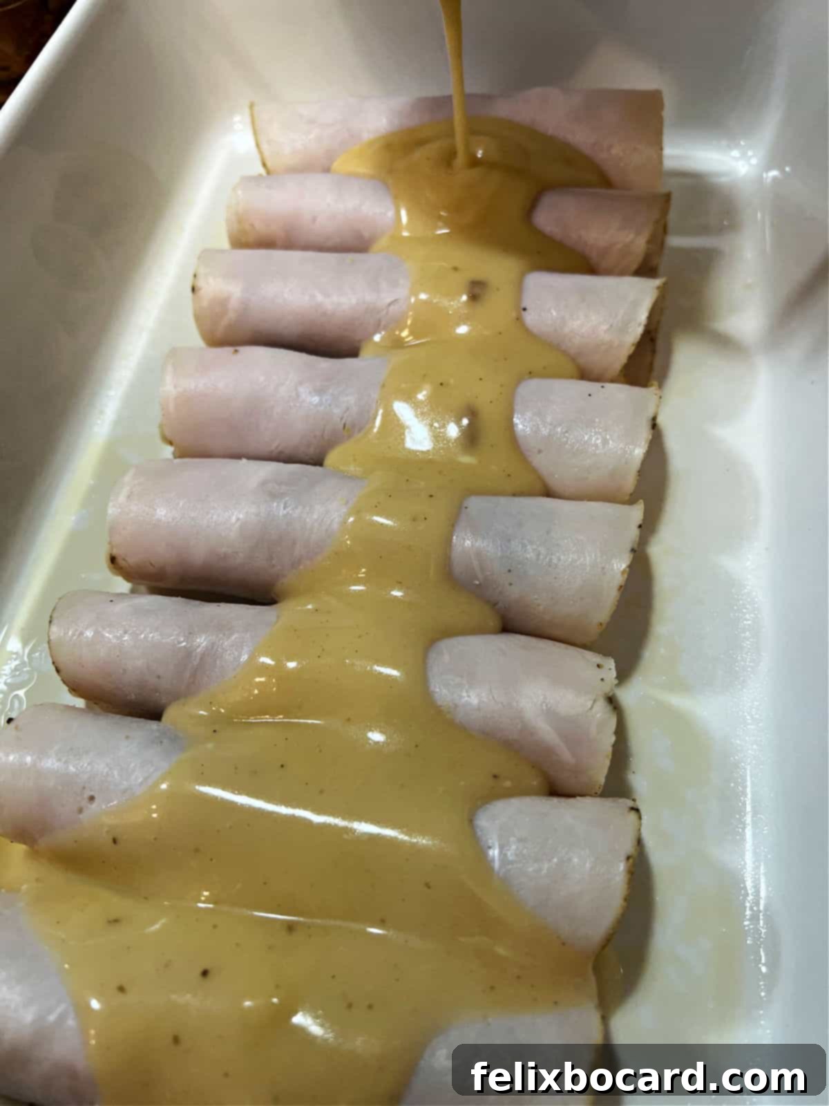 Thick, cheesy gravy being poured evenly over neatly arranged turkey roll-ups in a baking pan, prior to baking.