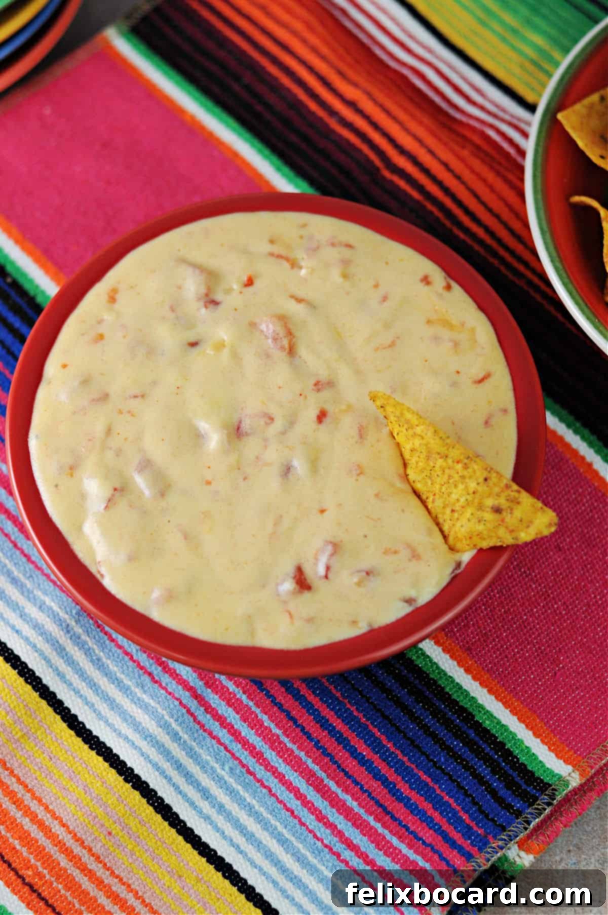 A vibrant bowl of homemade queso dip, perfectly melted and ready to serve, sitting on a colorful striped tablecloth, surrounded by tortilla chips.