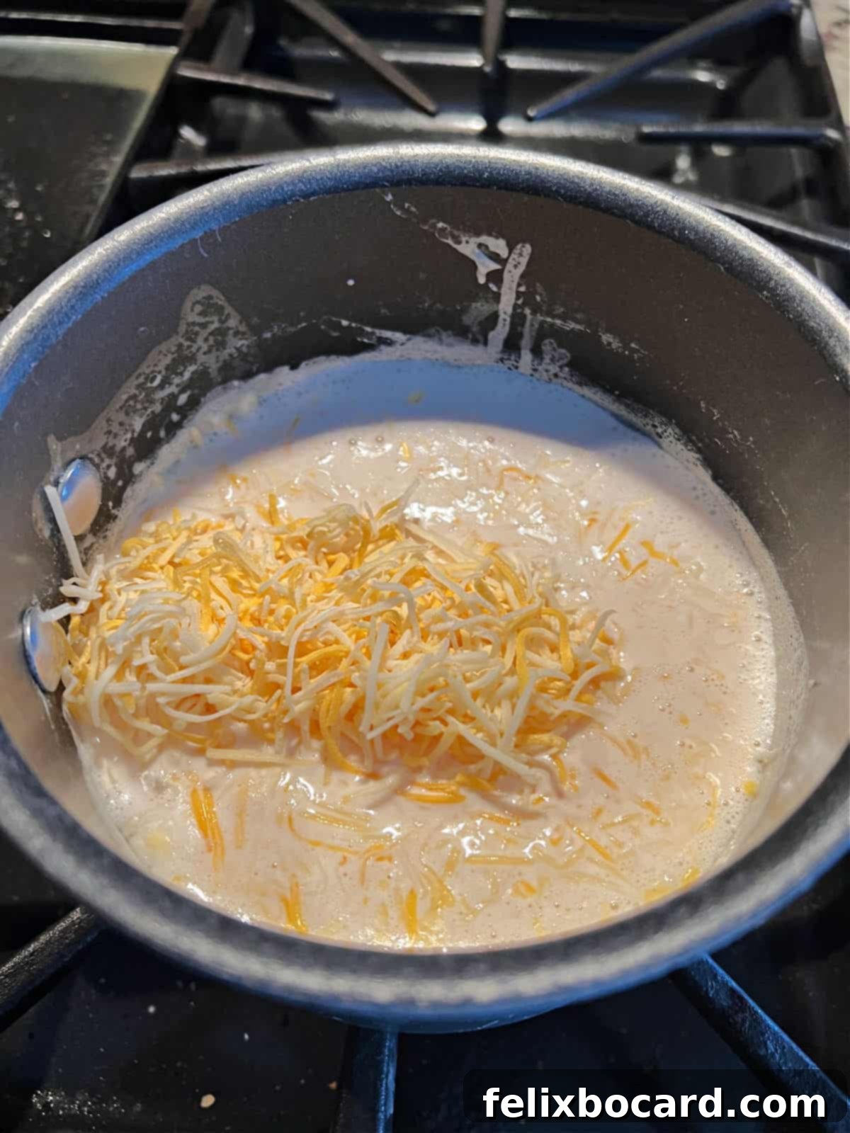 Close-up of shredded Mexican blend cheese being added to a saucepan filled with simmering evaporated milk, with a whisk stirring the mixture.
