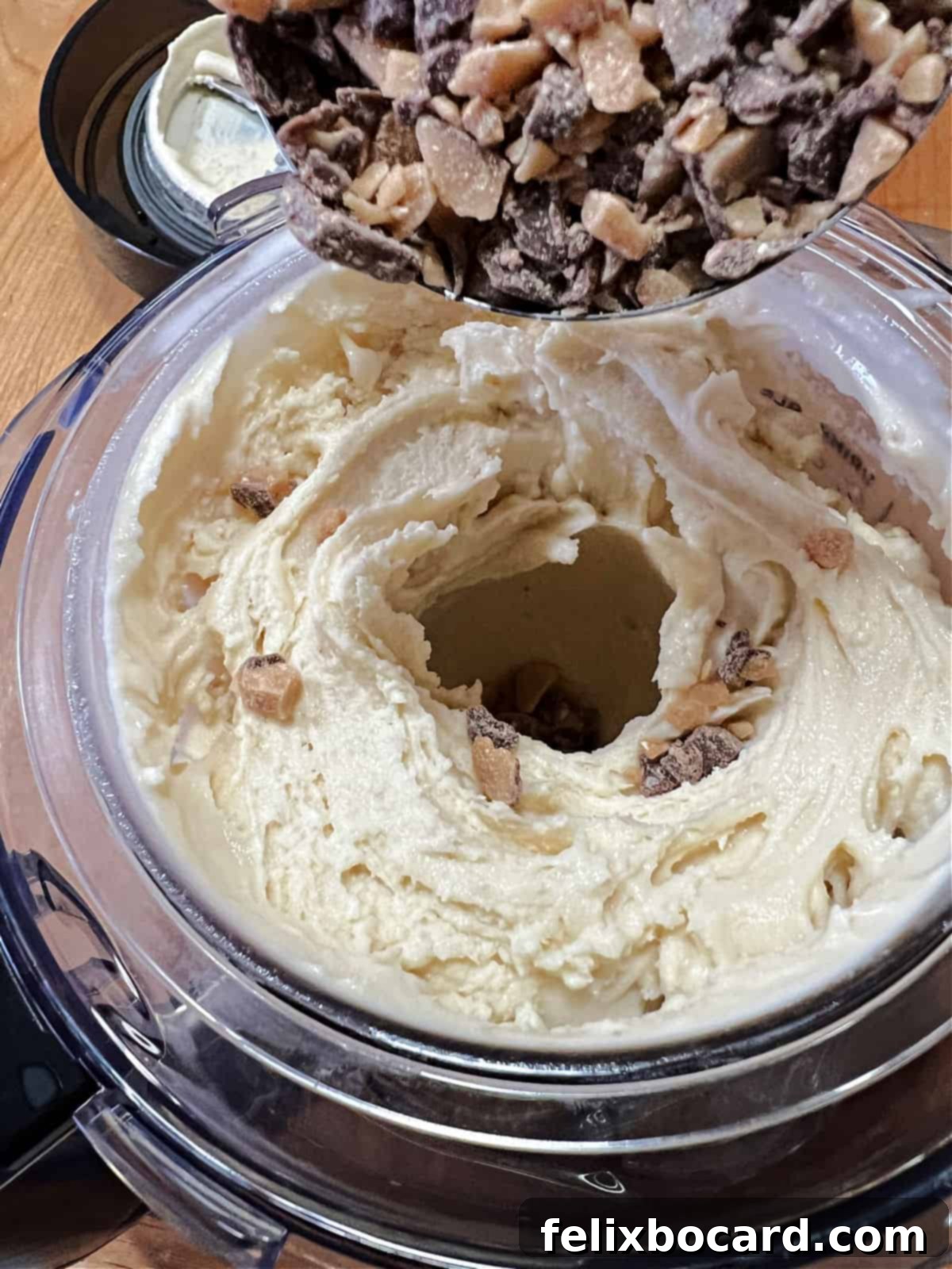 A spoon creating a deep well in the freshly churned ice cream, into which crunchy toffee bits are being added as a mix-in.