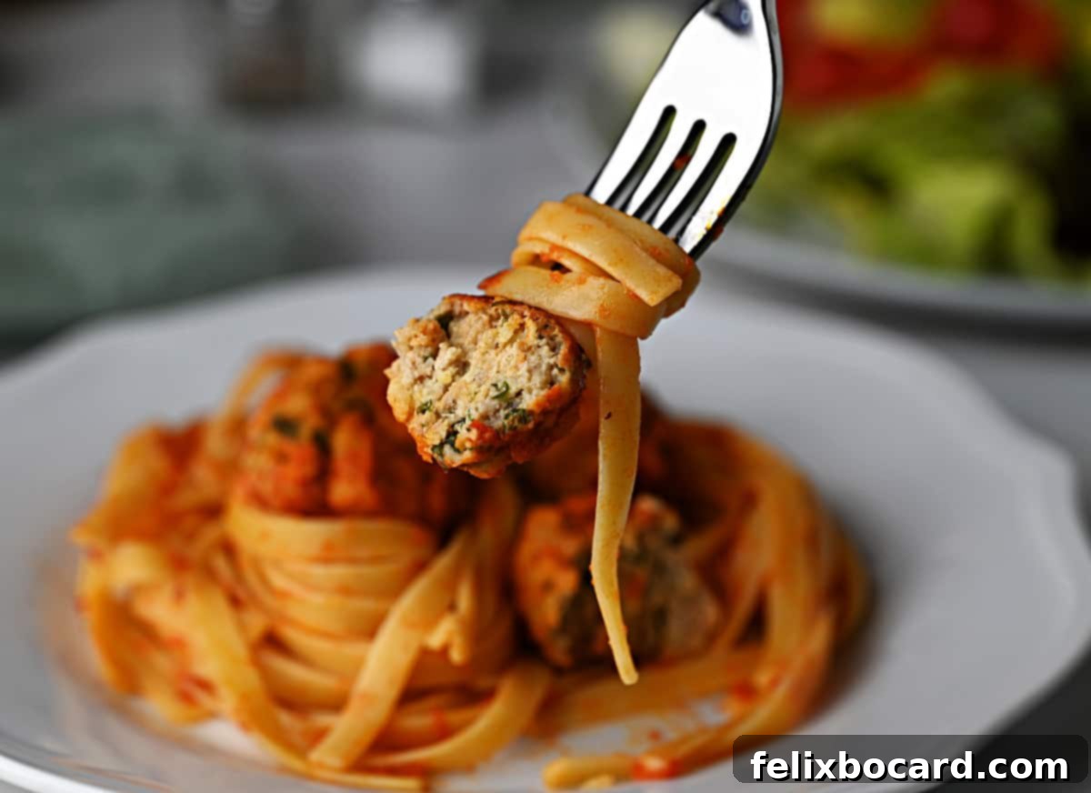 Fork holding a bitten chicken ricotta meatball with pasta and marinara sauce, garnished with fresh herbs.