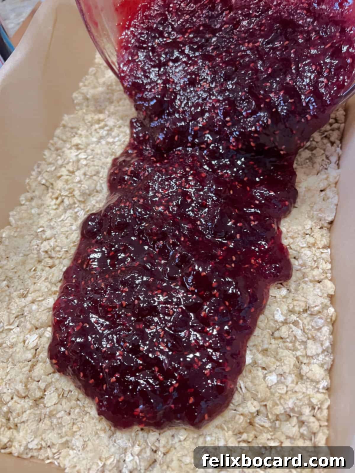 Pouring the raspberry jam mixture evenly over the oat crust in the baking pan.