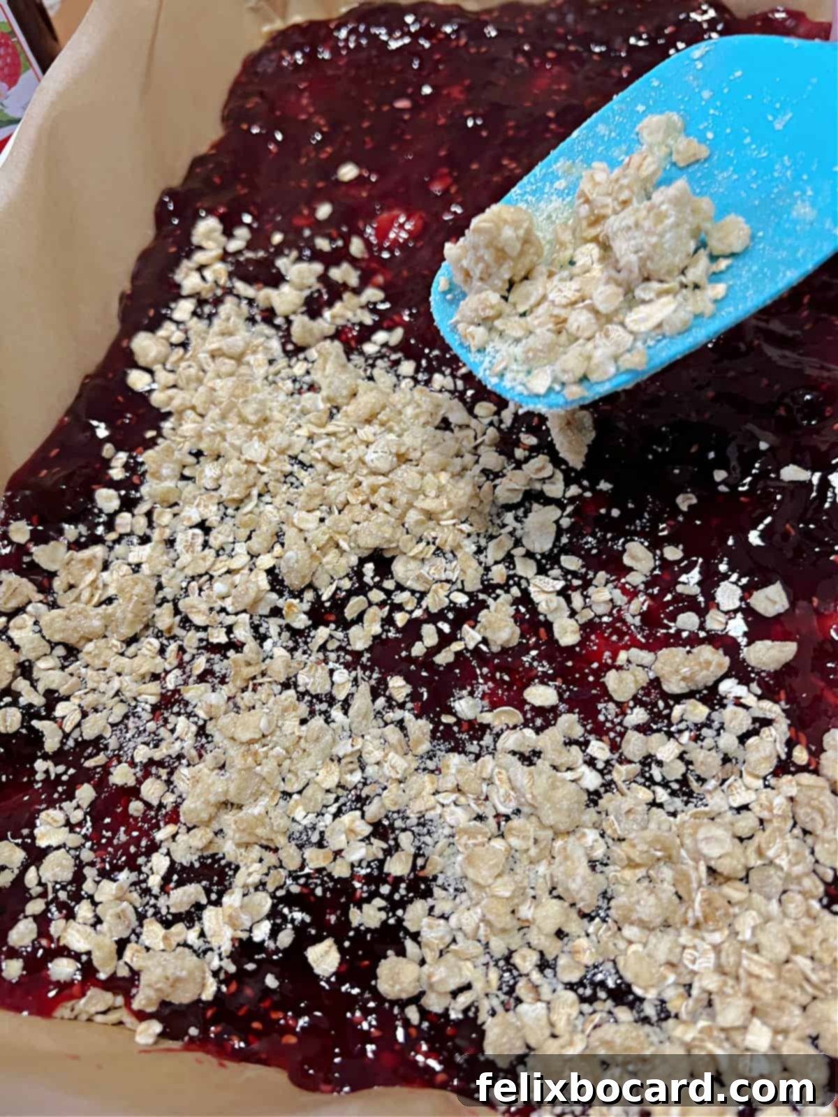 Sprinkling the remaining oat crumble mixture over the vibrant red jam layer in the baking pan.