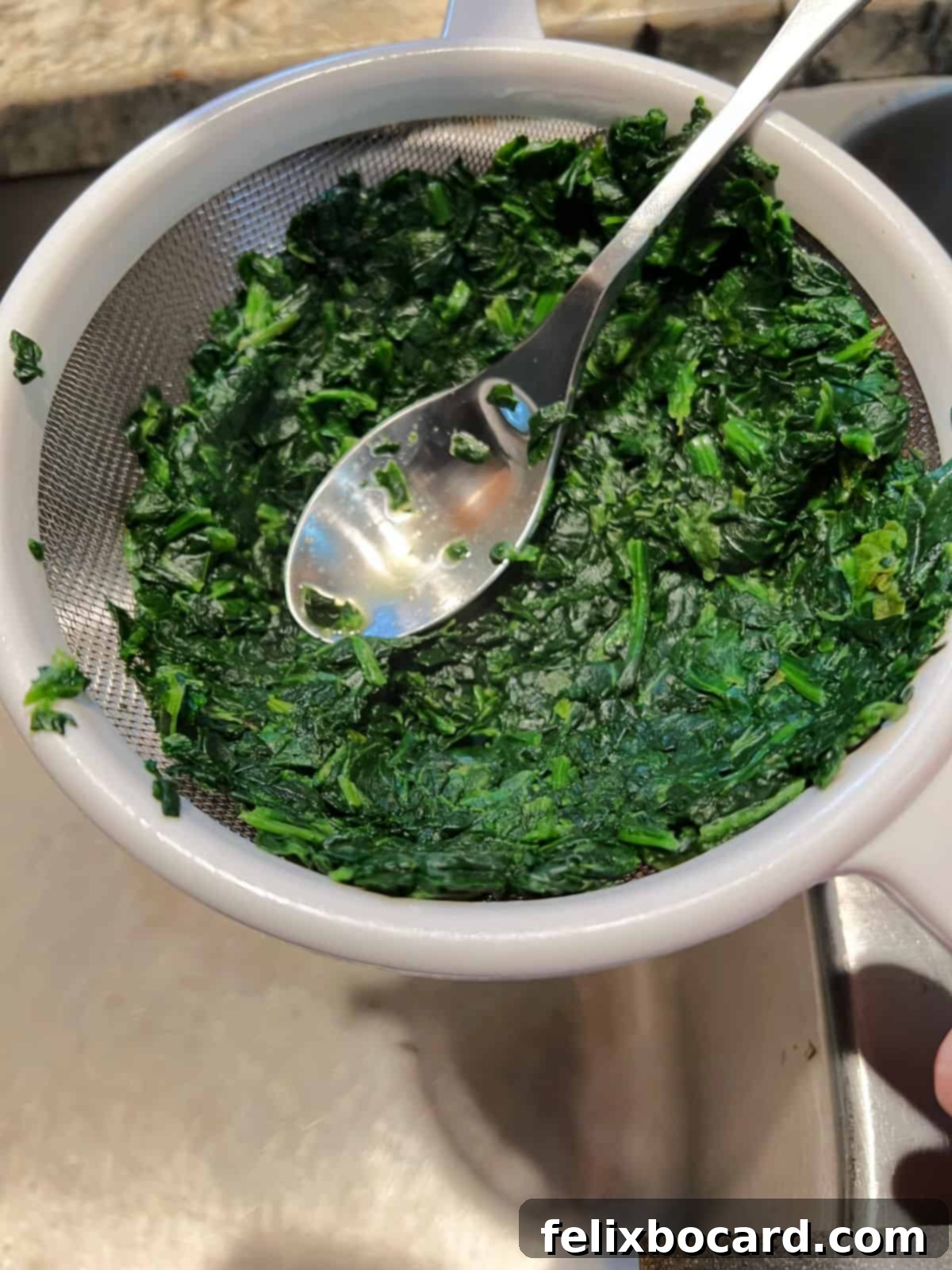 Squeezing excess water from microwaved spinach in a fine mesh strainer, ensuring a perfectly textured casserole.