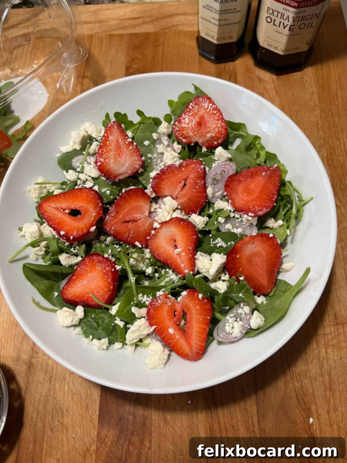 A partially assembled spinach and arugula salad in a bowl, showing mixed greens, strawberries, feta cheese, and sliced shallots before walnuts are added.