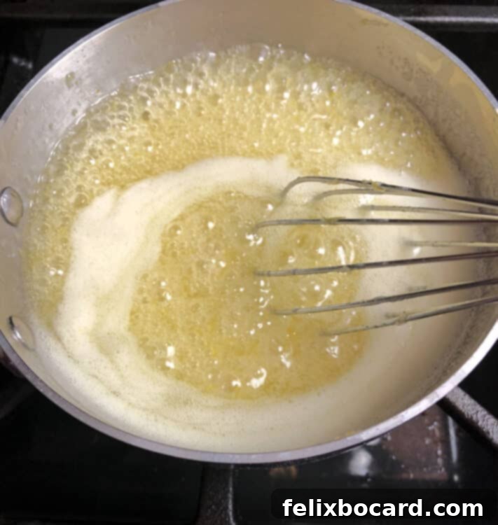 Close-up shot of the lemon curd filling being cooked in a small, gleaming non-reactive saucepan, with a whisk actively stirring the bright yellow mixture.