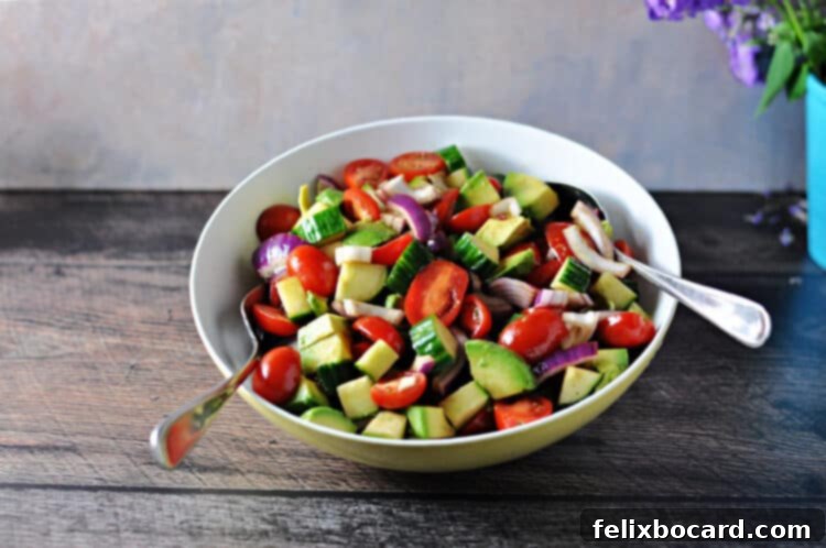 Balsamic Cucumber Tomato Salad served in a large white bowl with two silver serving spoons, showcasing vibrant red tomatoes, green cucumbers, and thinly sliced red onions.