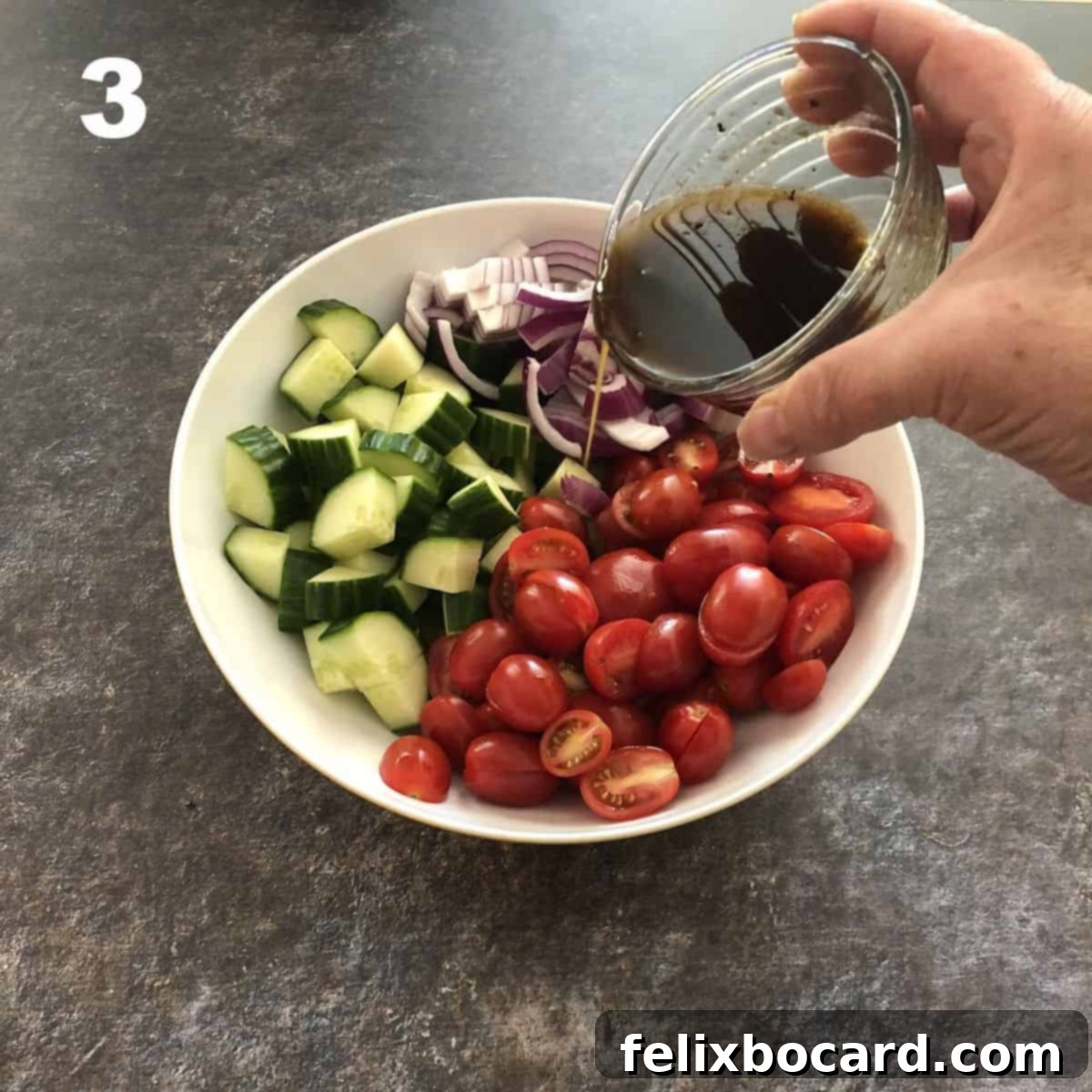 A stream of balsamic vinaigrette being poured from a small bowl into a larger bowl of chopped cucumbers, tomatoes, and red onions.