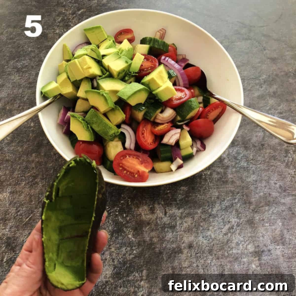 Diced avocado chunks being added to the prepared balsamic cucumber tomato salad in a bowl.