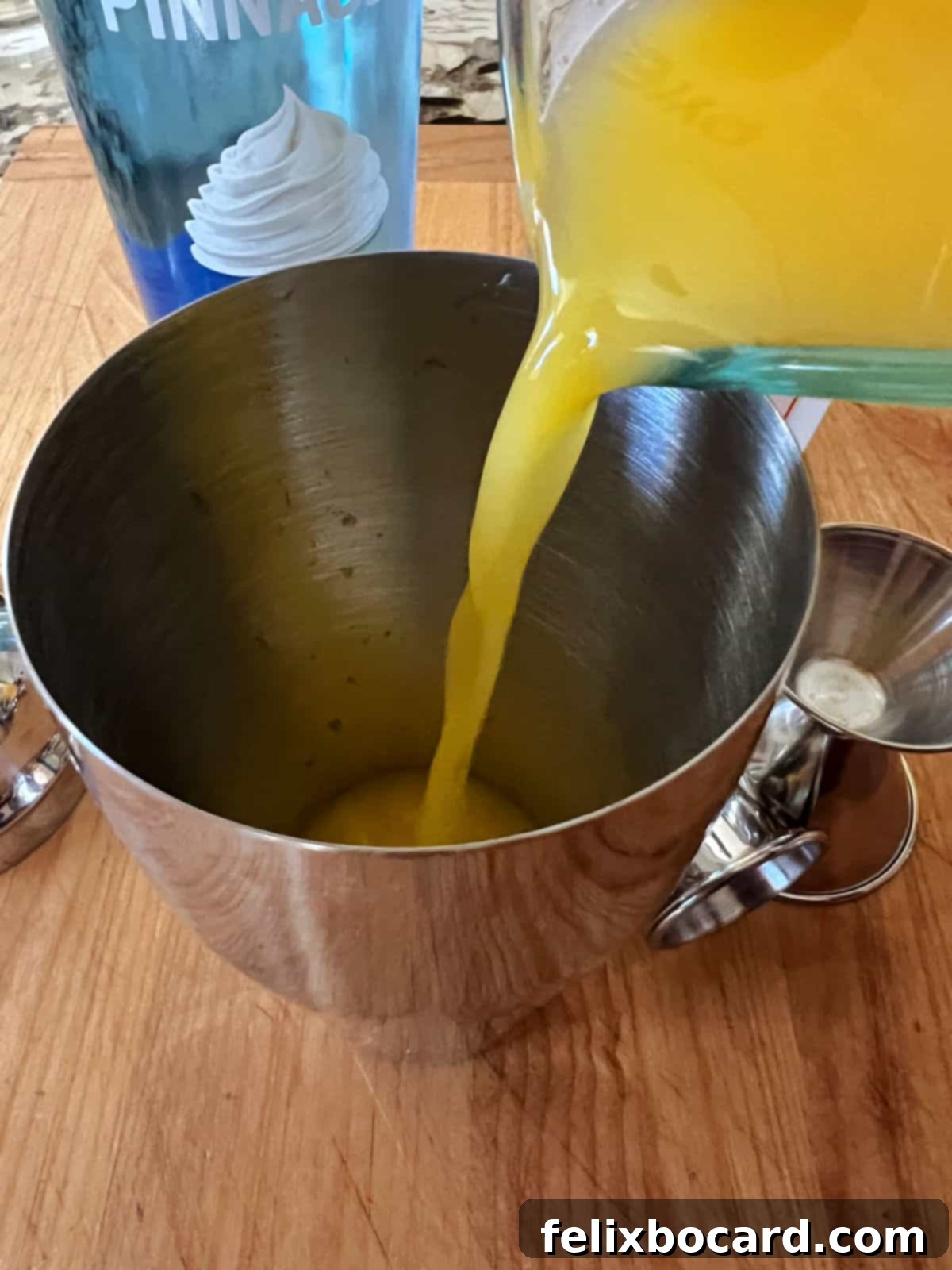 A close-up shot of orange juice being poured from a measuring cup into a sleek silver cocktail shaker filled with ice, capturing the preparation of the Creamsicle Cocktail.