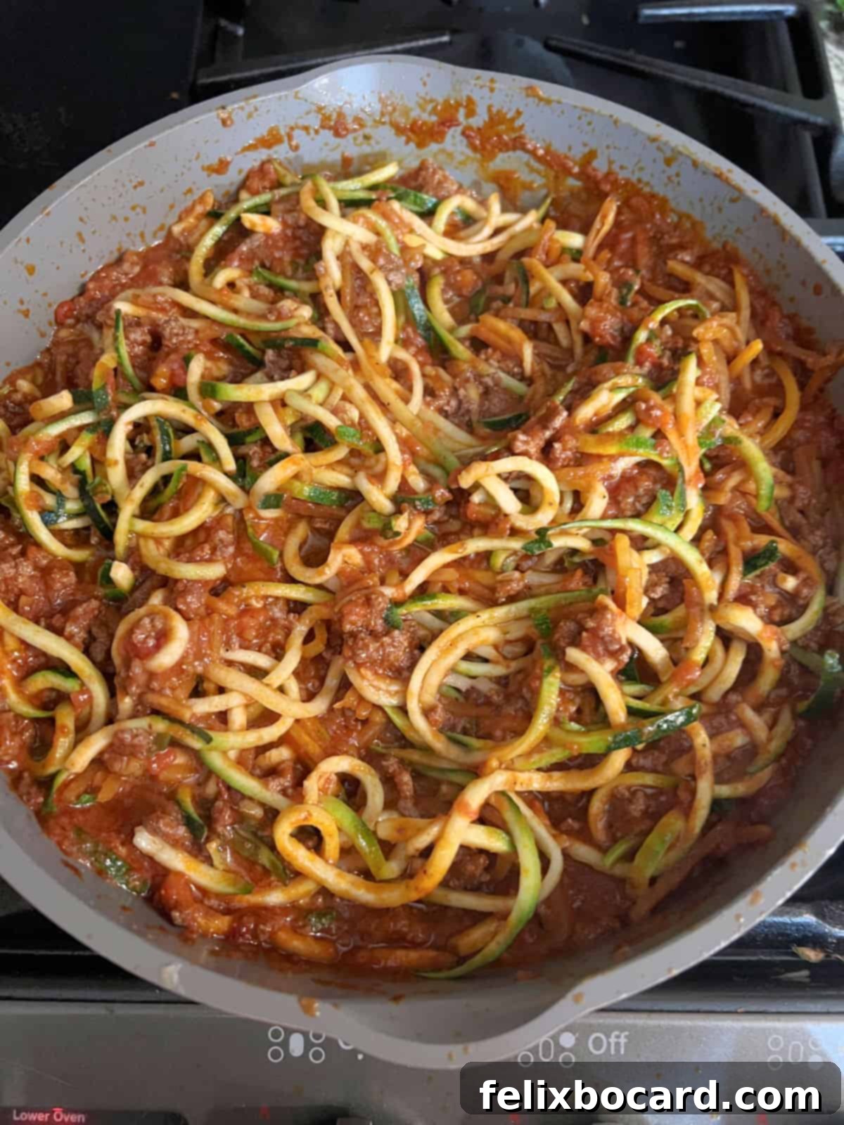 Spiralized zucchini noodles and butternut squash noodles being mixed into the spaghetti sauce in a large pan.