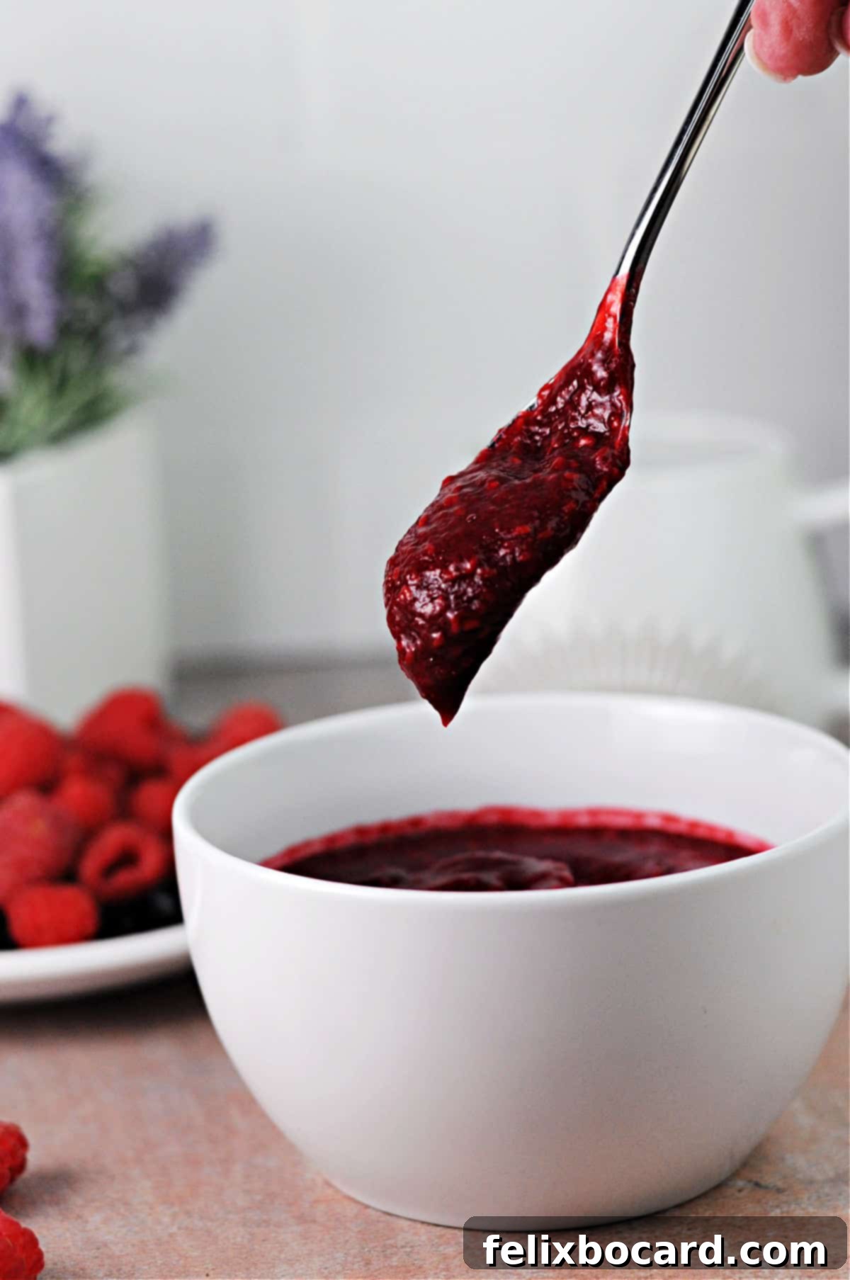 A spoon elegantly dripping rich, red keto raspberry sauce back into a ceramic bowl, showcasing its perfect consistency.