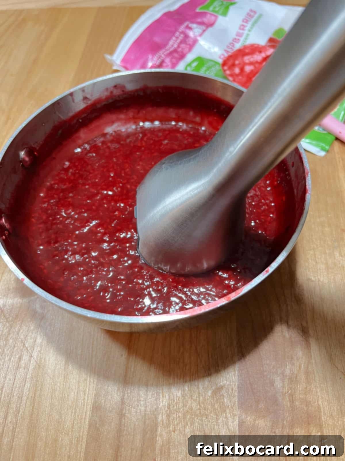 An immersion blender being used in a saucepan to make the cooked keto raspberry sauce smoother, reducing chunks and seeds.