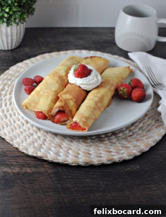 Three fruit-filled crepes beautifully arranged on a white plate with additional fresh fruit and a dollop of whipped cream, showcasing a delightful breakfast.