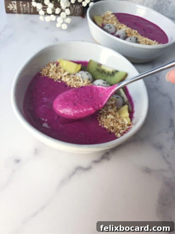 A spoon scooping up a vibrant magenta dragon fruit smoothie from a white bowl, adorned with toasted coconut and kiwi slices.