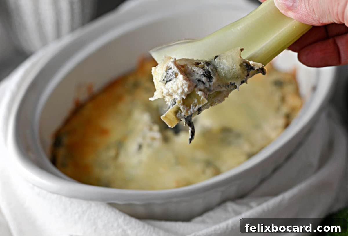 A hand holding a piece of celery with creamy keto spinach artichoke dip, showcasing its rich texture and delicious appeal.
