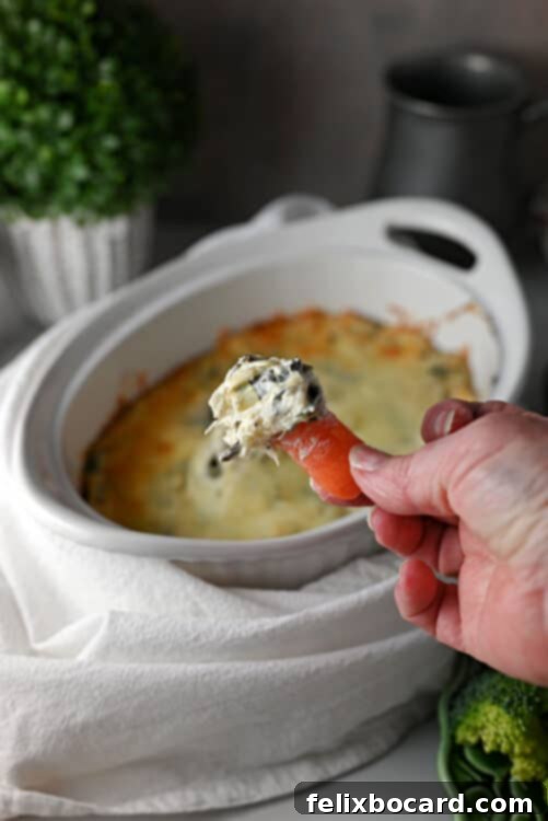 A hand dipping a baby carrot into a bowl of creamy spinach artichoke dip, highlighting its enticing texture.