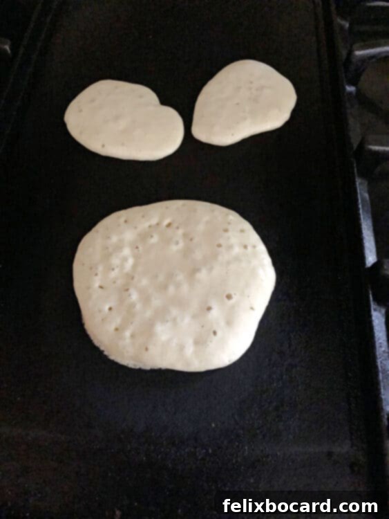 Pancake batter sizzling on a hot griddle, with one larger round pancake and two smaller oval-shaped pancakes cooking to a golden perfection, illustrating the two sizes needed for bunny butt pancakes.