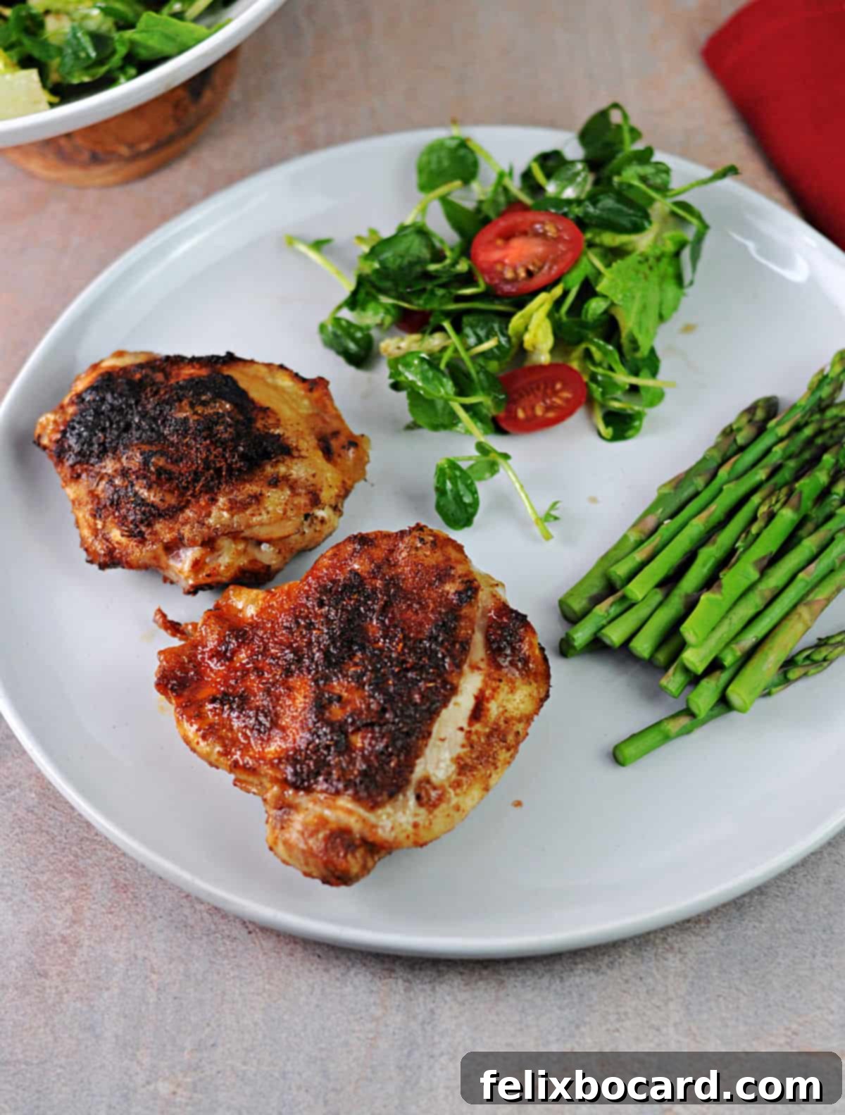Two air fryer cooked chicken thighs on a white plate, accompanied by a fresh green salad and roasted asparagus.