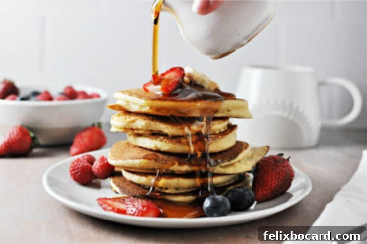 Stack of golden-brown oat milk pancakes topped with fresh mixed berries and rich maple syrup being poured over them.