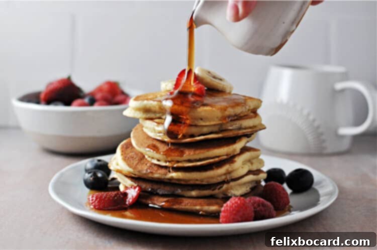Golden maple syrup being poured over a stack of fluffy oat milk pancakes, with a side of fresh fruit including berries and banana slices.